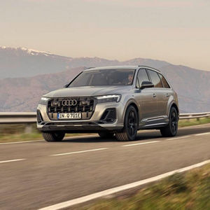 A silver Audi Q7 TFSI e drives on a winding road with mountains in the background, showcasing its modern design and elegance.