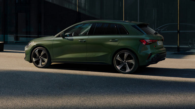 A green Audi A3 Sportback car parked beside glass buildings in sunlight.