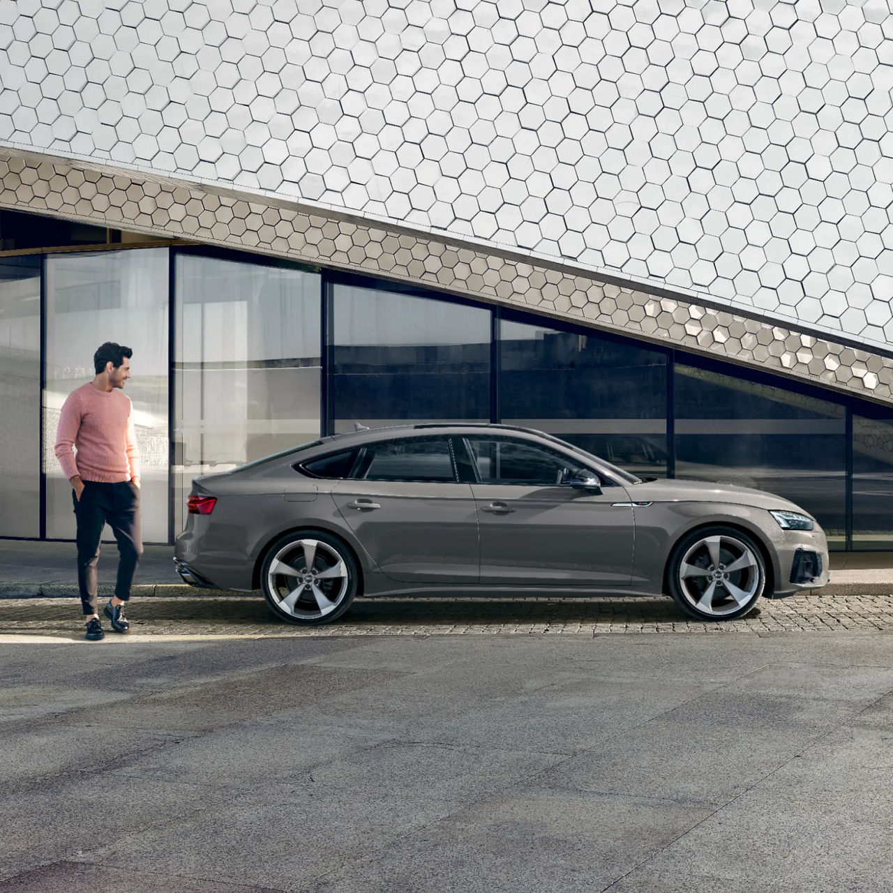 A person standing by a grey sedan in front of a modern building with hexagonal pattern facade.