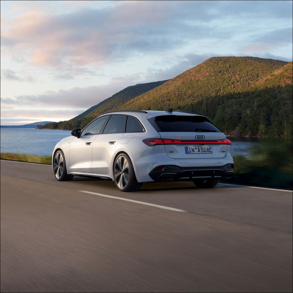 A sleek white Audi car drives along a scenic road by a lake with lush green hills in the background under a partly cloudy sky.