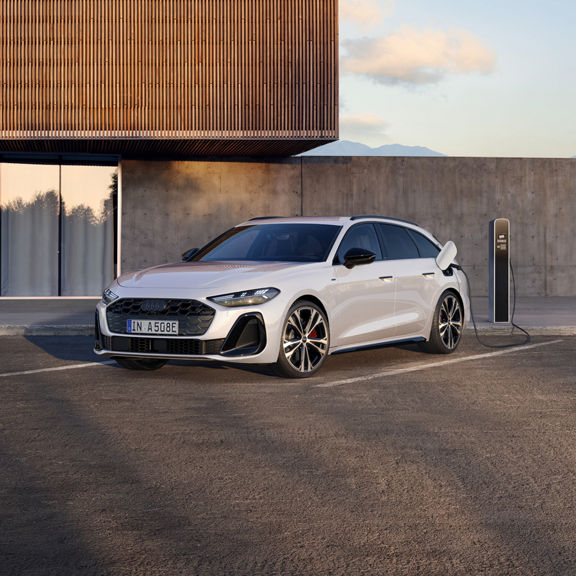A sleek white Audi e-hybrid car is parked and charging at a modern station beside a concrete wall and a wooden-paneled building.