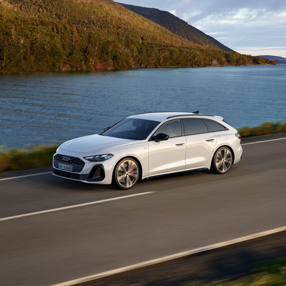 Sleek white Audi car driving on a scenic lakeside road, flanked by lush greenery under a clear blue sky.