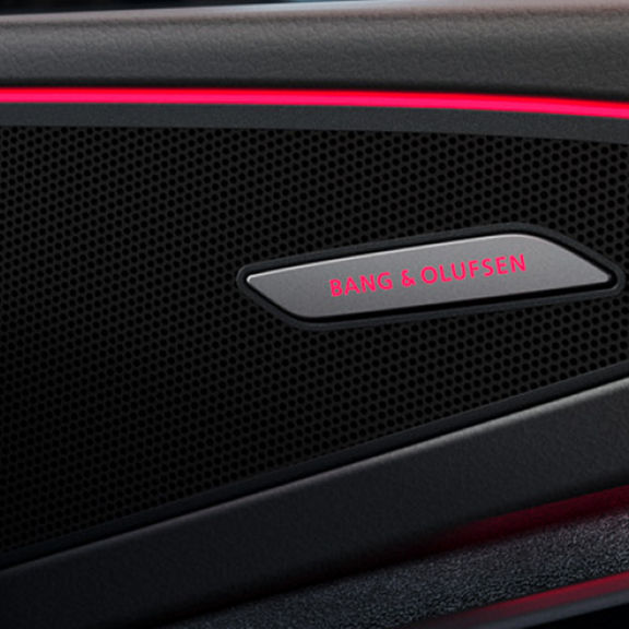 Close-up of a Bang & Olufsen speaker on a car door, featuring sleek black mesh and subtle red ambient lighting.