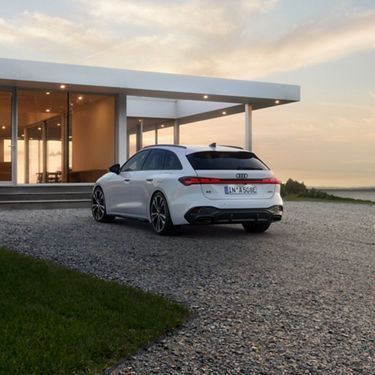 A sleek white car is parked on a gravel driveway beside a modern glass house. The setting sun casts a warm glow, creating a serene and elegant atmosphere.