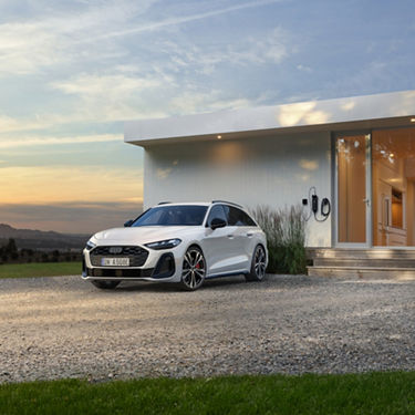 A sleek white Audi parked beside a modern house at sunset. The vehicle is positioned on a gravel driveway, adjacent to a wall-mounted electric charger.