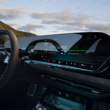 Car dashboard with a sleek digital display showing drive modes and battery charge. Steering wheel and scenic sky with clouds visible outside.
