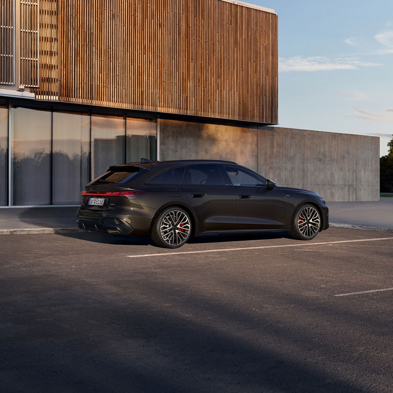 A sleek black car is parked in front of a modern building with wooden slats. The setting sun casts a warm glow, highlighting the car's stylish design.