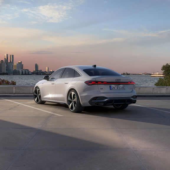 A sleek white car is parked on a waterside pavement at sunset, with a city skyline in the background and a ferry on the water to the right.