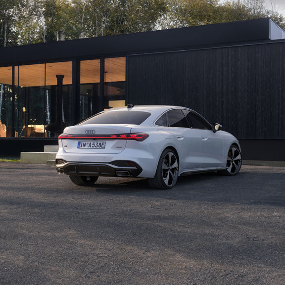 A sleek white sedan parked on a driveway beside a modern glass house, surrounded by greenery and trees.
