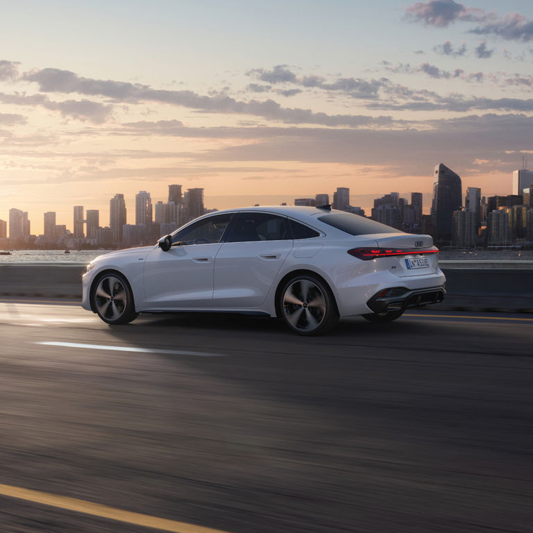 A sleek white car drives along a highway at sunset, with a modern city skyline in the background.