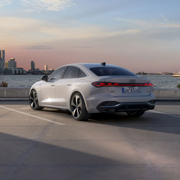 A sleek white car parked on a waterfront lot during sunset. The city skyline and a passing ferry are in the background.