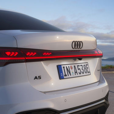 Rear view of a white Audi A5 with sleek lines and illuminated taillights at dusk. The license plate reads "IN A538E." Calm ocean in the background.