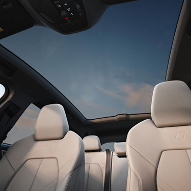 Car interior showing beige leather seats with embossed "S" logo and a panoramic sunroof overlooking a blue sky with soft clouds.