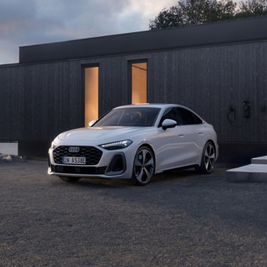 A sleek white sedan is parked beside a modern, dark-paneled building at dusk.