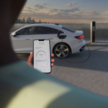 A person holds a smartphone showing an app displaying an electric car's battery status. The car is charging at a station by the water, with a city skyline under a cloudy sky.