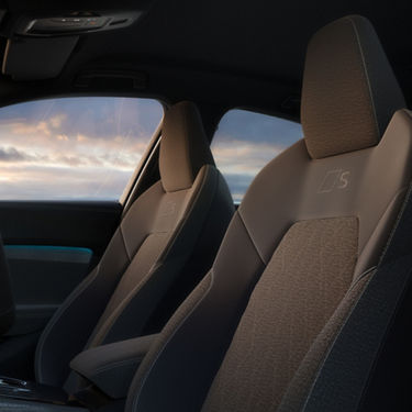 Front seats of a car with textured gray upholstery, featuring an embossed "S" logo. The window shows a serene sky with soft, pastel clouds.