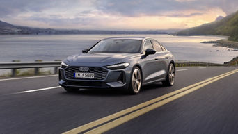 Sleek grey car on a scenic coastal road during sunset. The sea and hills are in the background.