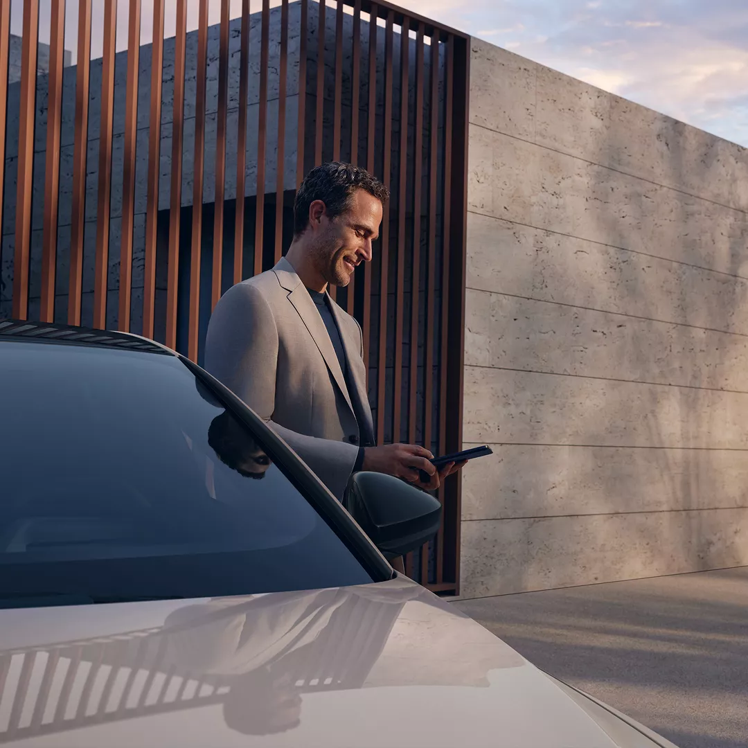 A man in a suit stands next to a sleek car, holding a phone, in front of a modern architectural backdrop.