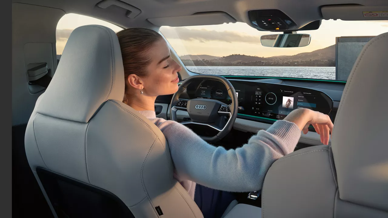 Relaxed driver enjoys the sleek Audi EV interior with digital dash and scenic waterfront view.
