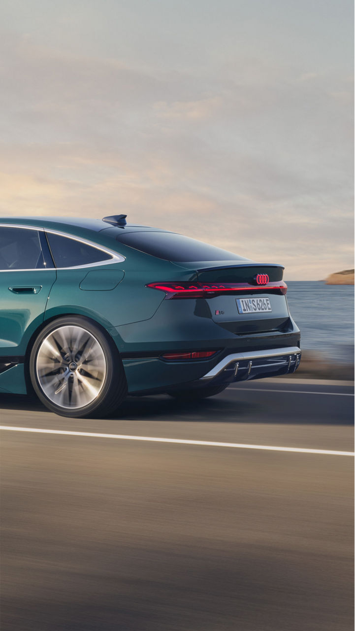 A sleek green car drives along a coastal road, with mountains and water in the background, illuminated by a serene sunset.