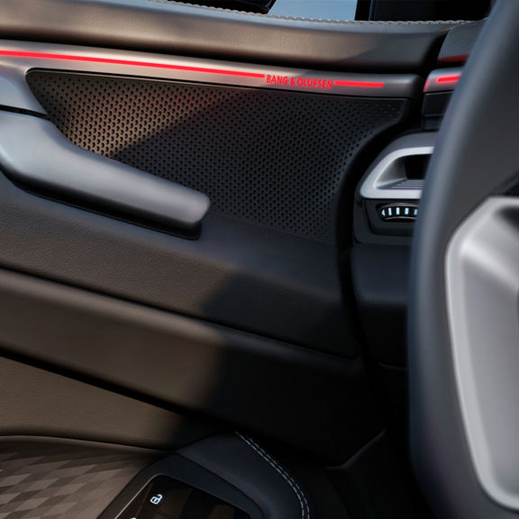Close-up of a car door interior featuring a Bang & Olufsen speaker, sleek black leather, and illuminated red accents.