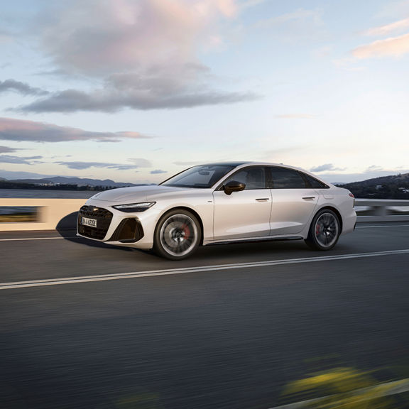 A silver Audi A6 Saloon e-hybrid drives along a coastal road at sunset, showcasing its elegant design and performance.