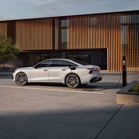 A white Audi A6 Saloon e-hybrid car is plugged into a charging station outside a modern building, surrounded by greenery and sunlit concrete.