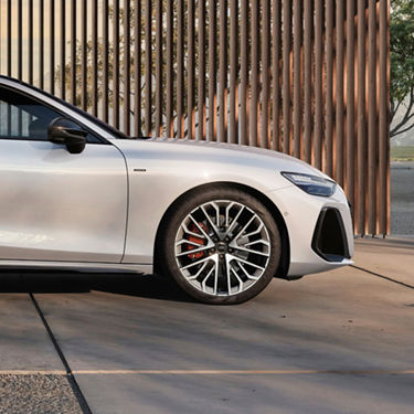 A silver car parked beside wooden slats, showcasing its sporty design and distinctive alloy wheels.