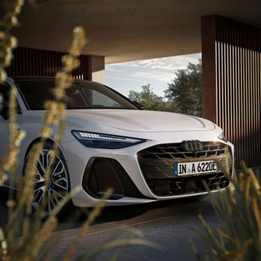 A white Audi sports car with distinctive headlights is parked under a modern structure, surrounded by greenery.