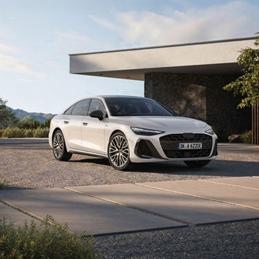A white Audi is parked in a modern driveway, surrounded by greenery and a contemporary home, under a clear sky.