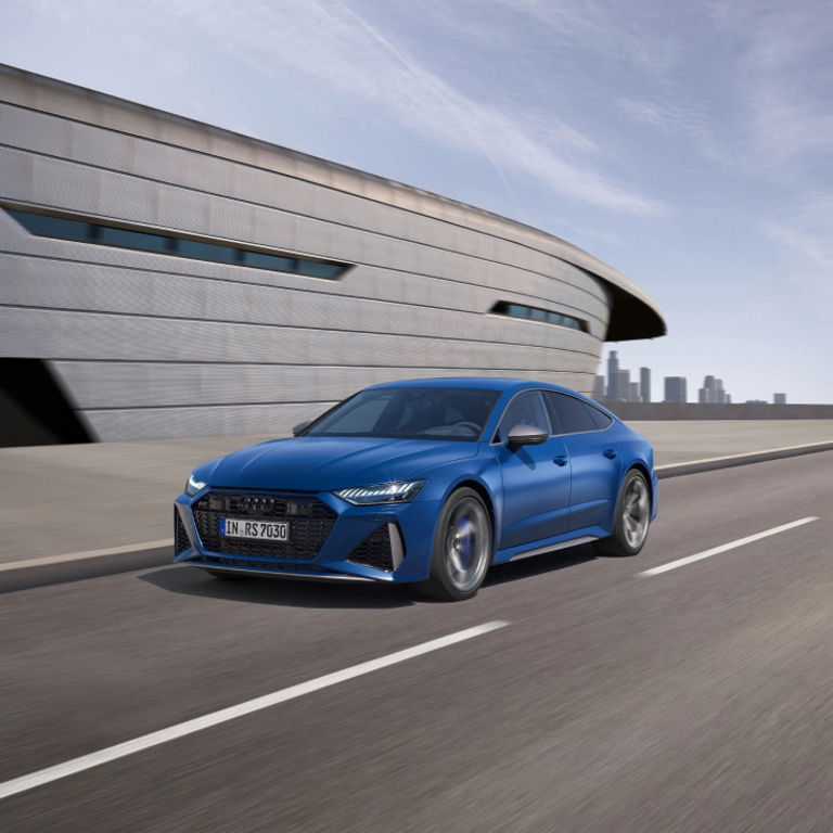 Blue sedan driving on road with modern building and city skyline in the background.