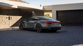 A sleek gray Audi RS e-tron GT charging outside a modern home, with elegant architecture and a textured stone wall in the background.