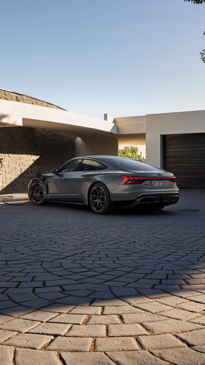 A sleek gray Audi RS e-tron GT charging outside a modern home, with elegant architecture and a textured stone wall in the background.