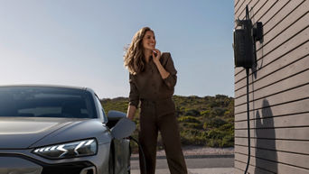 A woman in a brown outfit stands next to a grey electric car, connected to a charging station on a modern building.