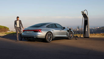 A silver Audi S e-tron GT is parked beside a modern EV charging station, with a city skyline in the background.