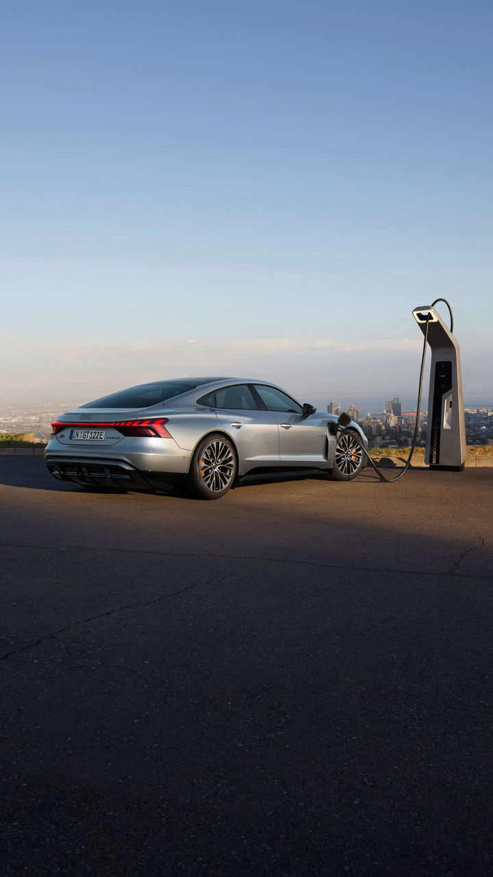 A silver Audi S e-tron GT is charging at a sleek station with a scenic city view in the background during a clear sky.