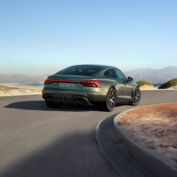 n Audi e-tron GT Performance sports car rounds a curve on a scenic road with mountains and ocean in the background, showcasing its dynamic design.
