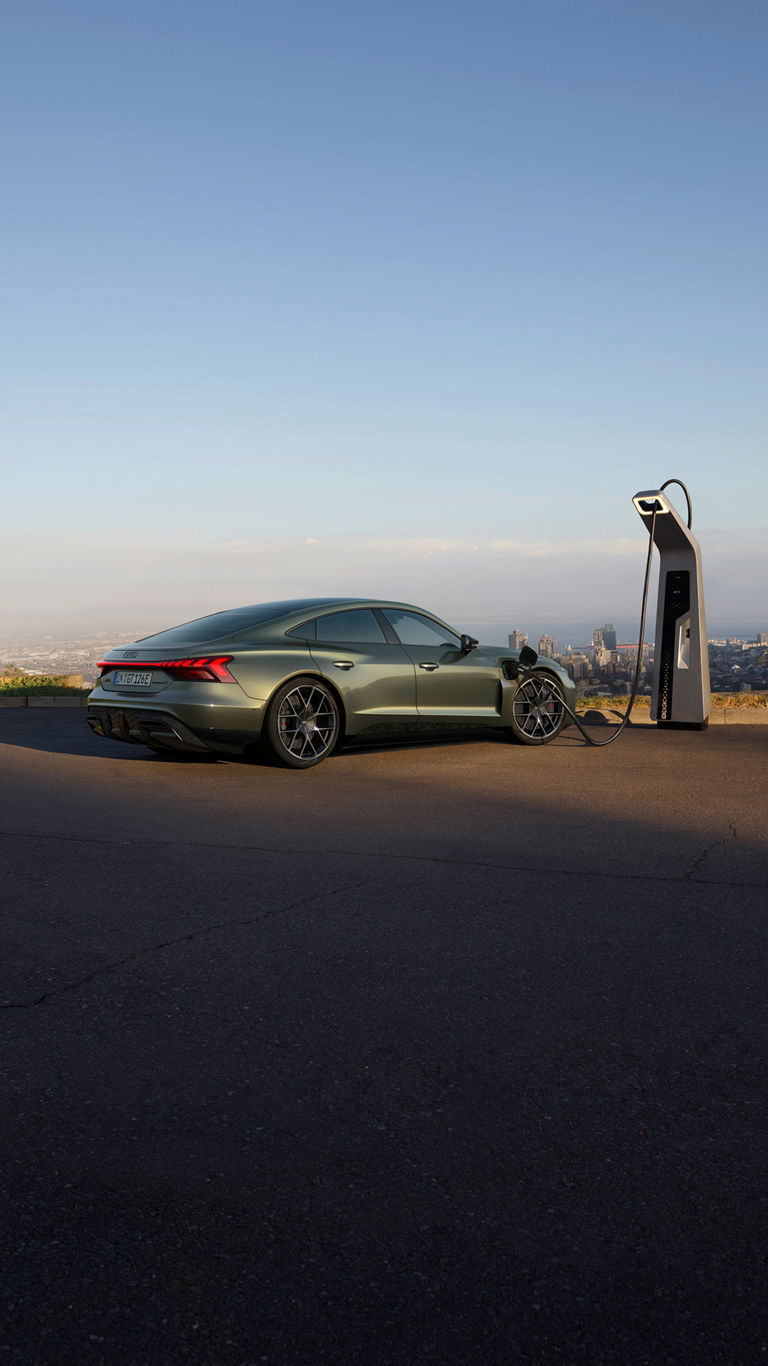 A green Audi RS e-tron GT car charging at a modern station with a city skyline in the background under a clear sky.