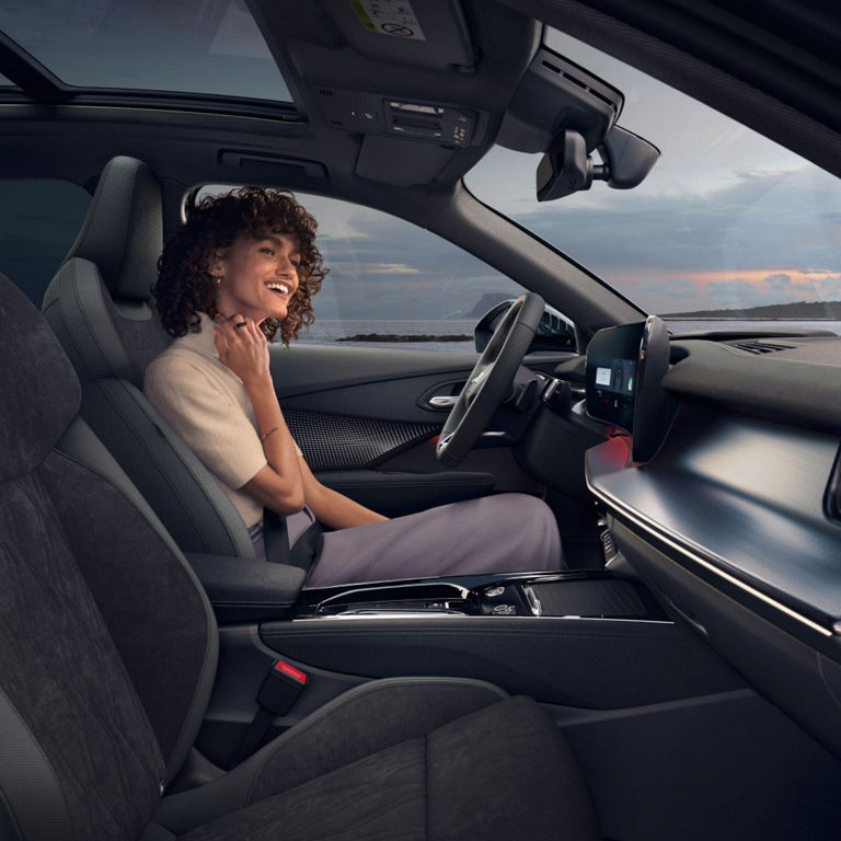 A woman sits in a modern car's front seat, with a scenic view visible through the large windshield, showcasing a stylish interior design.
