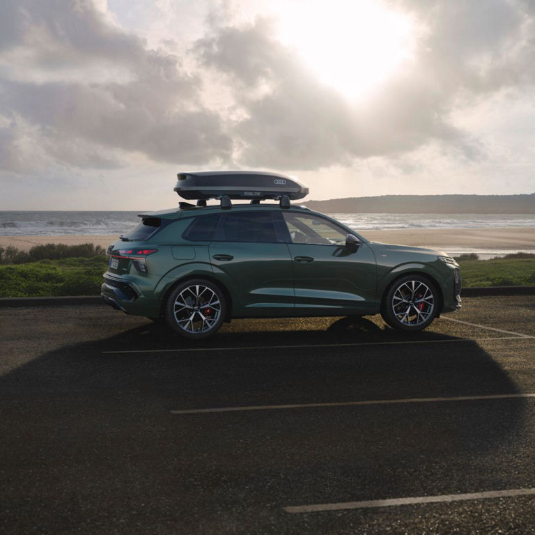 A green Audi SUV parked along a coastal road, featuring a rooftop cargo box, with the sun peeking through clouds in the background.