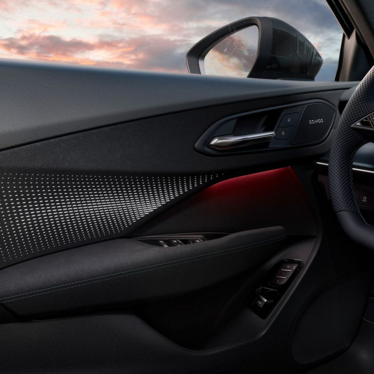 Close-up of a car door interior featuring ambient lighting, speaker, and control buttons against a backdrop of a colourful sunset.