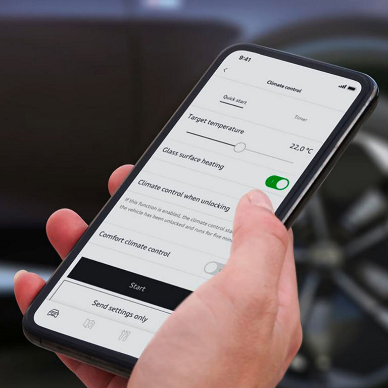 A hand holds a smartphone displaying car climate control settings, including target temperature and glass surface heating options.