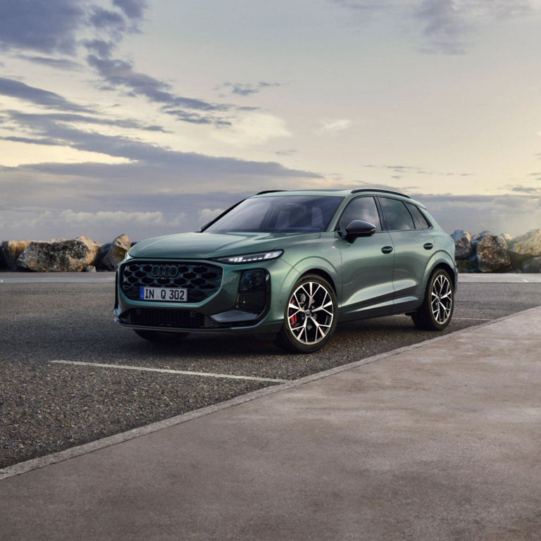 A green Audi SUV parked on a coastal road, with rocky shores and a cloudy sky in the background.