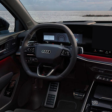 Interior of an Audi showcasing a modern steering wheel, digital dashboard, and sleek controls against a scenic seaside backdrop.