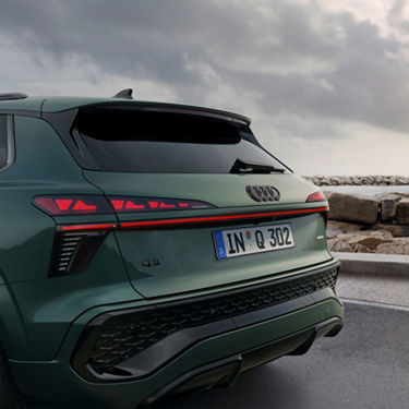 A rear view of an Audi Q3 SUV in a sleek green finish, showcasing modern taillights and a coastal backdrop under a cloudy sky.