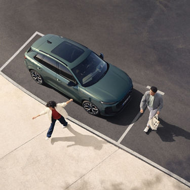 A green SUV is parked on a concrete lot, while a person joyfully dances nearby and another walks towards the vehicle with a bag.