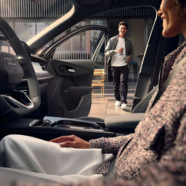 A woman sits inside an Audi, looking towards a man outside holding a drink, against a modern architectural backdrop.