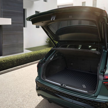 The open trunk of a sleek green SUV reveals a spacious cargo area with a textured mat, set against a modern home backdrop.