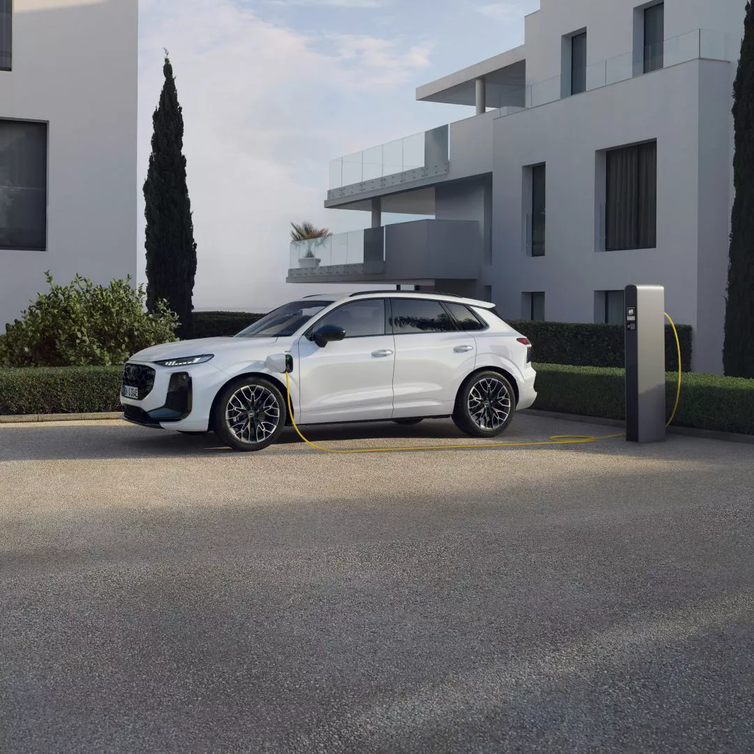 A white Audi Q3 e-hybrid plugged in an EV charger while parked in front of modern houses