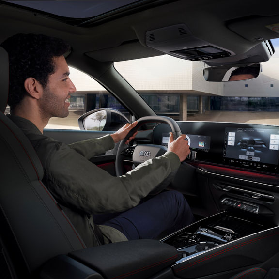 A person driving an Audi Q3, showing the modern dashboard, steering wheel, and touchscreen controls.